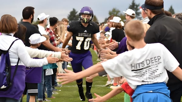 Estudiantes de la Portland Christian High School se reúnen en uniformes deportivos en un campo mientras los espectadores están de pie al fondo.
