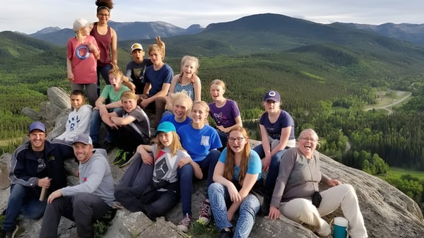 Un grupo de estudiantes y adultos de la Prairie Christian Academy están sobre una roca mirando hacia montañas boscosas.