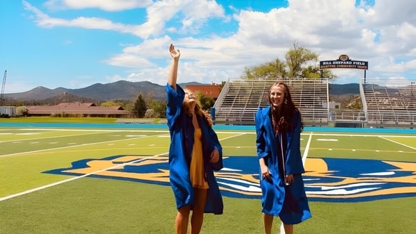 Dos personas en togas de graduación azules están en el campo deportivo del Prescott Unified School District frente a montañas y un cielo nublado.