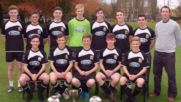 Un grupo de estudiantes del Presentation Brothers College posan juntos en un campo de hierba con árboles y edificios de fondo.