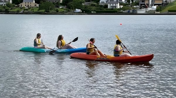 Alumnos de la Presentation Secondary School (Cork) están remando en kayaks inflables en un cuerpo de agua con la ciudad de fondo.