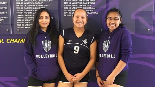 Tres estudiantes de la Queen Elizabeth High School están en sus uniformes de voleibol frente a una pared con premios deportivos.