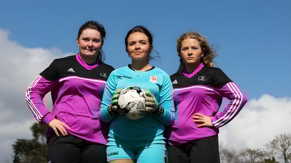 Tres futbolistas en uniforme con un balón están frente a un cielo azul en el terreno de Queen Elizabeth's 6th Form.