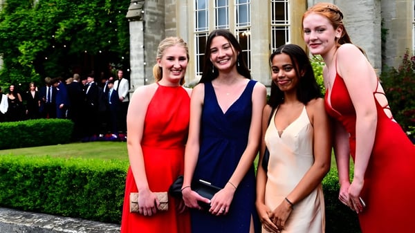 Cuatro estudiantes de Queen’s College están juntas en un jardín vestidas de manera formal con un edificio de fondo.