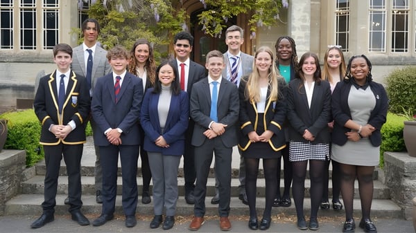 Un grupo de estudiantes en ropa formal posan en la escalera frente a un edificio en el campus de Queen’s College.