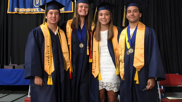 Cuatro estudiantes de la Queensbury High School están juntos en togas de graduación en un escenario con un banner azul y amarillo de fondo.