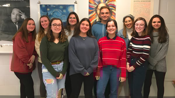 Un grupo de estudiantes está en el aula del Queensbury Union Free School District frente a coloridos carteles en la pared.