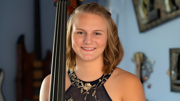 Una joven sostiene un violín frente a una pared con obras de arte enmarcadas en el Queensbury Union Free School District.
