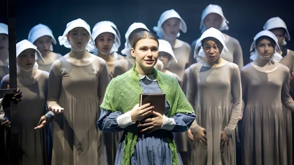 Una mujer con bufanda verde sostiene un libro frente a un grupo de mujeres en túnicas blancas en el campus de la Queenswood School.