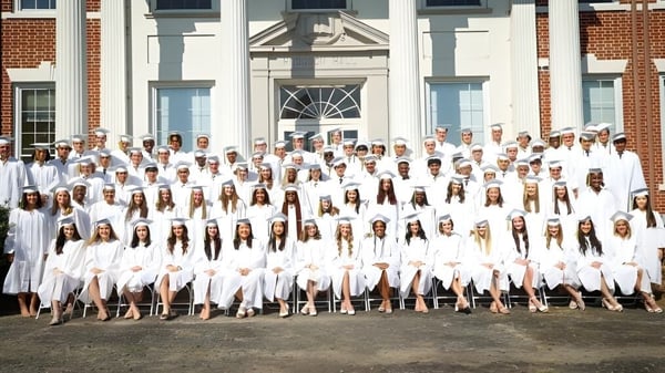 Estudiantes de la Rabun Gap-Nacoochee School en vestidos de graduación blancos se reúnen frente a un edificio histórico de ladrillo.