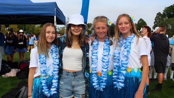 Un grupo de alumnas de la Rangiora High School lleva coloridas guirnaldas de flores frente a una carpa azul en un evento al aire libre.