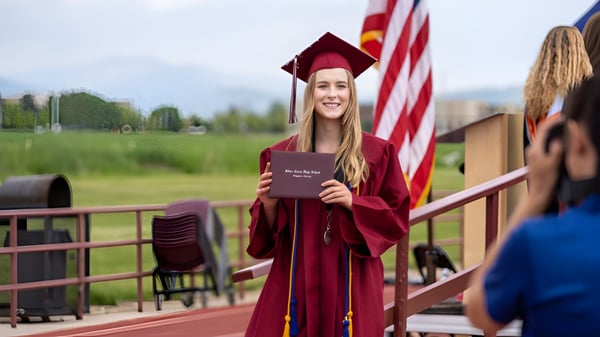 Una joven en toga de graduación roja está en el balcón de la Red Creek High School con vista a montañas y árboles.