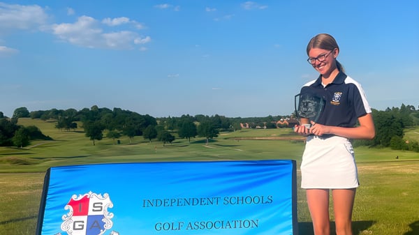Una estudiante de Reddam House Berkshire está frente a un cartel de la Independent Schools Golf Association con un campo de golf y un bosque al fondo.