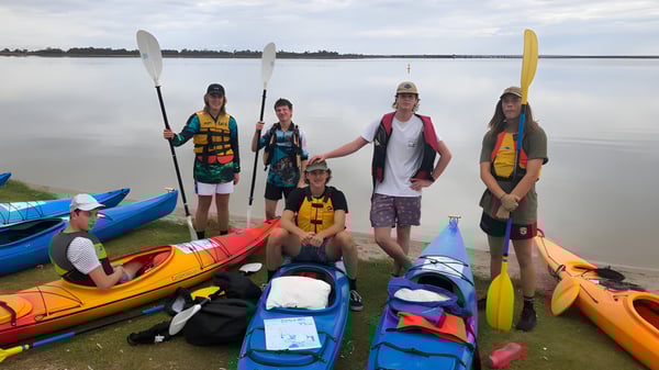 Alumnos de la Renmark High School están en la orilla del lago entre coloridos kayaks y paddleboards en un paisaje tranquilo.