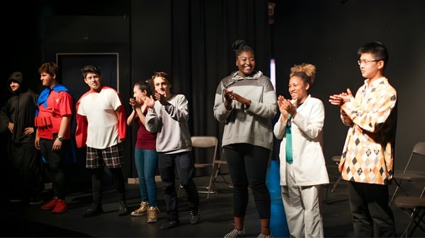 Estudiantes de Ribet Academy están en un escenario en una sala débilmente iluminada con cortinas e imágenes de fondo.