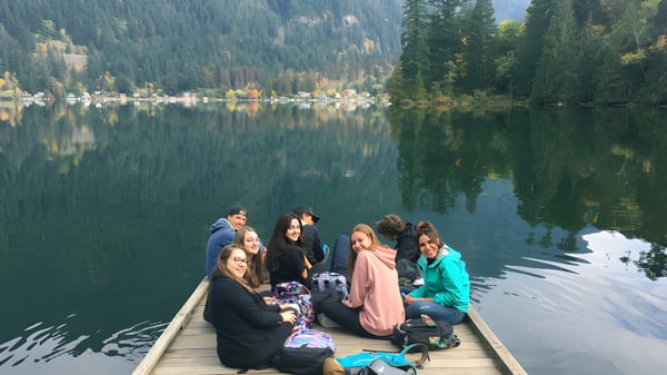 Un grupo de estudiantes está sentado en un muelle de madera junto a un lago con montañas boscosas al fondo en el campus de la Rick Hansen Secondary School.