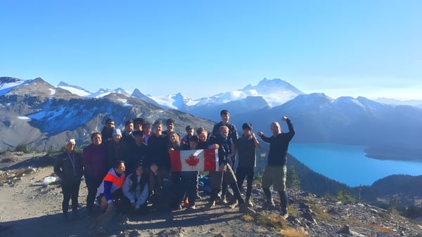 Un grupo de estudiantes de la Rick Hansen Secondary School está en una cresta montañosa con vistas a cumbres nevadas y un lago.