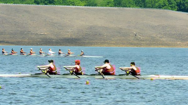 El equipo de remo del Rivermont Collegiate entrena en un cuerpo de agua con una orilla cubierta de hierba y árboles al fondo.