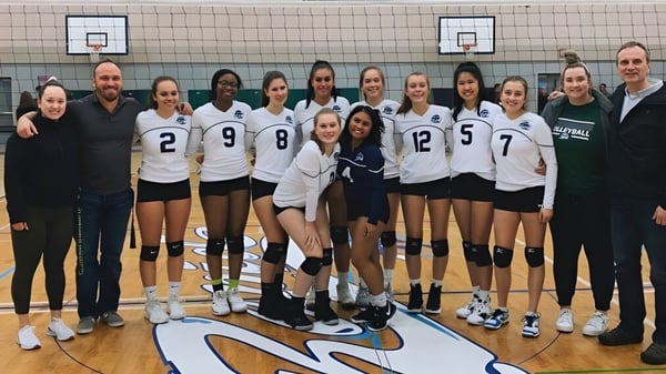 Estudiantes de la Riverside Secondary School están en sus uniformes de voleibol en una cancha de baloncesto con canastas al fondo.