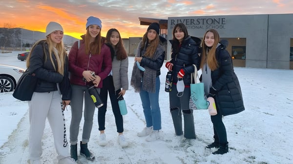 Un grupo de alumnas está afuera en la nieve frente a la Riverstone International School con un colorido atardecer de fondo.