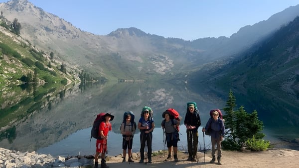Un grupo de estudiantes está en la orilla rocosa de un lago frente a altas montañas en el terreno de la Riverstone International School.