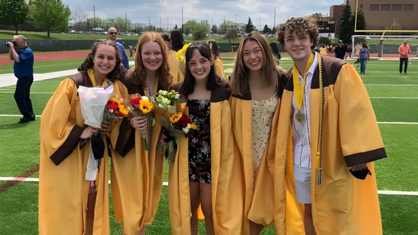 Un grupo de jóvenes mujeres en ropa de graduación sostiene flores en el campo deportivo de Rochester Community Schools.