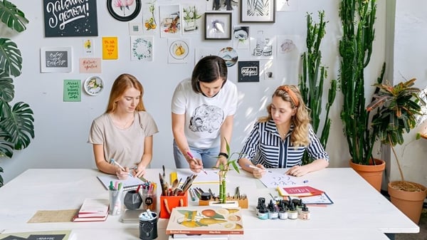 Tres estudiantes de la Rockwell Charter School están sentados en una mesa trabajando en dibujos y pinturas en la clase creativa.