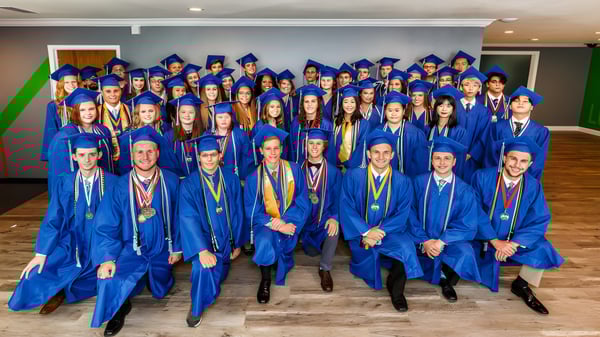 Un gran grupo de graduadas y graduados en togas azules están en el salón de Rocky Bayou Christian High School.