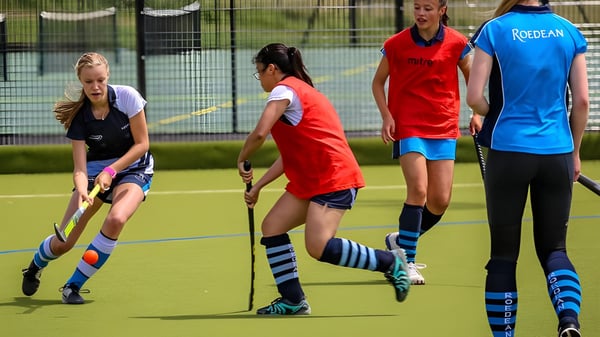Alumnas de la Roedean School juegan al hockey sobre césped en un campo de césped artificial con una cerca y árboles al fondo.