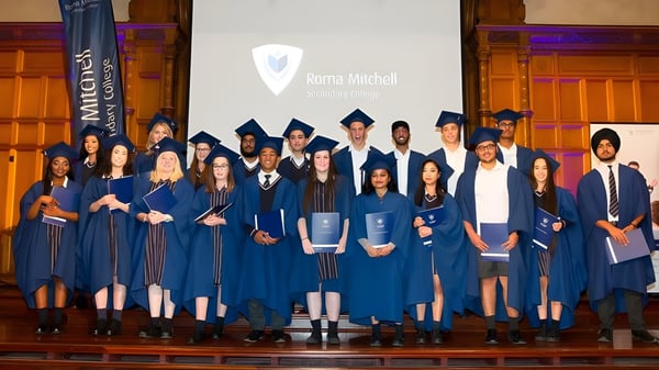 Los graduados en togas azules están en el escenario frente a una pantalla con el logo del Roma Mitchell Secondary College.