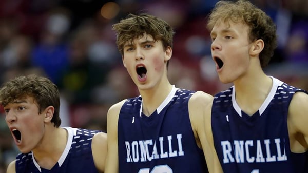 Tres estudiantes de la Roncalli Catholic High School en uniforme de baloncesto durante el juego con expresiones faciales intensas.