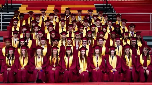 Estudiantes de la Roncalli Catholic High School están juntos en un escenario con togas rojas y doradas durante la ceremonia de graduación.
