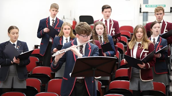 Un grupo de alumnos en uniformes de la Royal School está sentado con partituras en una sala de conferencias con sillas rojas.
