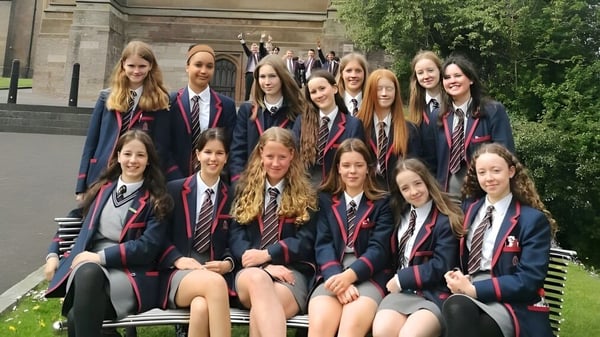 Un grupo de jóvenes alumnas en uniforme escolar están sentadas en un banco frente a un edificio histórico de la Royal School Armagh.