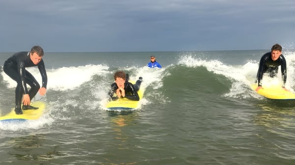Estudiantes de la Royal School Cavan surfean con trajes de neopreno en olas en el océano bajo un cielo nublado.
