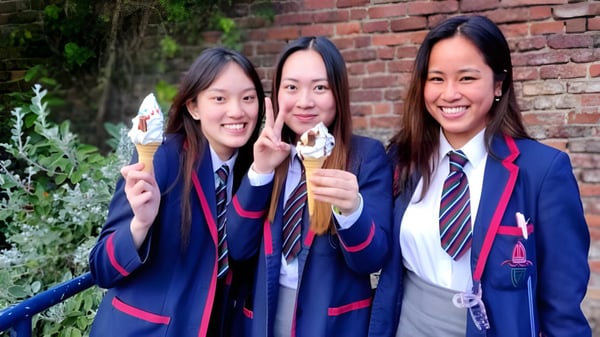 Tres estudiantes de la Royal School en uniformes escolares sostienen helados y están en un jardín frente a una pared de ladrillo.