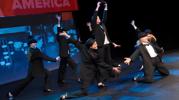 Un grupo de estudiantes de la Rugby School baila y gesticula en un escenario con la palabra America de fondo.