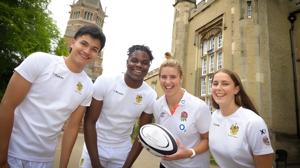 Un grupo de cuatro estudiantes en camisetas deportivas blancas está frente a un edificio histórico con una alta torre en el campus de la Rugby School.