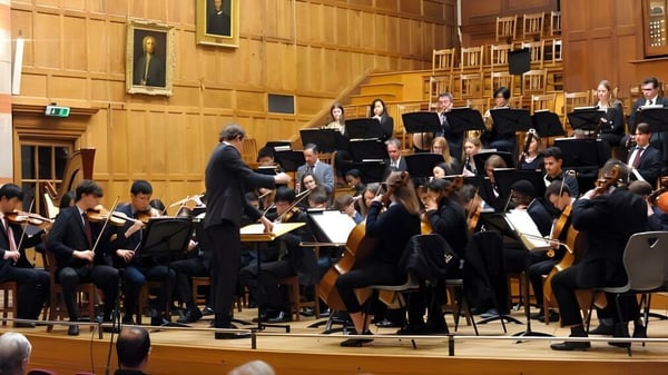 La orquesta de la Rugby School toca en un escenario en una gran sala de conciertos con paneles de madera.
