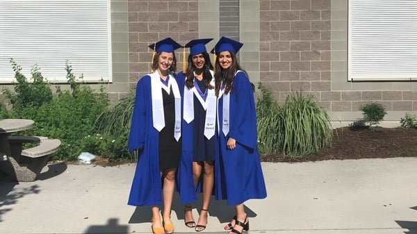Tres estudiantes de la Rutland Senior Secondary School están de pie en túnicas de graduación azules frente a un edificio de ladrillo.