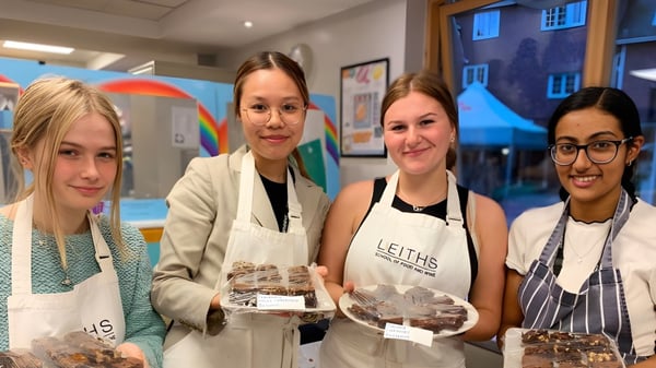 Cuatro estudiantes de la Rye St. Antony School llevan delantales y presentan productos horneados en una colorida cocina escolar.