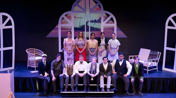 Alumnos y alumnas del Sacred Heart College están de pie en ropa formal frente a un escenario colorido con ventanales y detalles arquitectónicos.