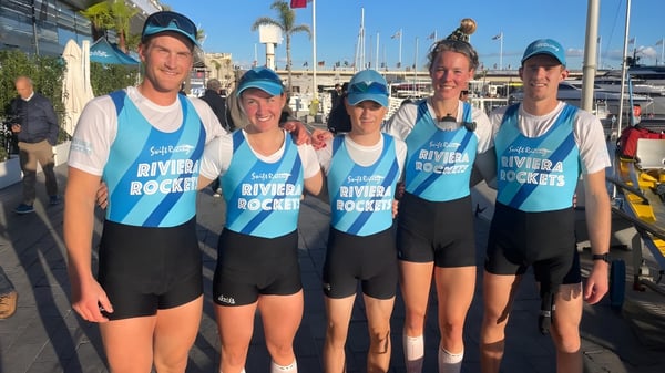 Un grupo de estudiantes del Sacred Heart College está en uniformes azules y blancos frente a un puerto con barcos y edificios.