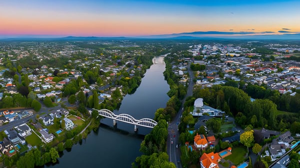 Toma aérea de una ciudad junto a un río con montañas verdes al fondo durante el atardecer.