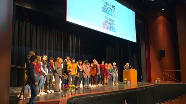 Estudiantes de la Sacred Heart High School están en el escenario frente a una gran pantalla de proyección con texto.