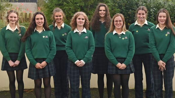 Un grupo de alumnas de la Sacred Heart School está de pie en el uniforme escolar verde afuera en un prado frente a árboles y un edificio.