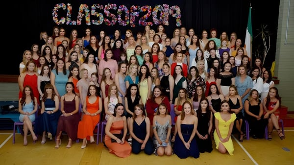 Un gran grupo de alumnas de la Sacred Heart School posan con vestidos coloridos en un gimnasio frente al letrero CLASSOF2020.