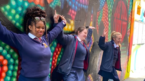 Un grupo de alumnas y alumnos de la Sacred Heart Secondary School en uniforme escolar está frente a un colorido mural y levantan los brazos.