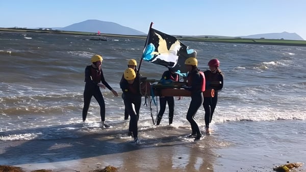 Alumnos de la Sacred Heart Secondary School en trajes de neopreno están en el agua poco profunda cerca de una playa con un paisaje montañoso de fondo.
