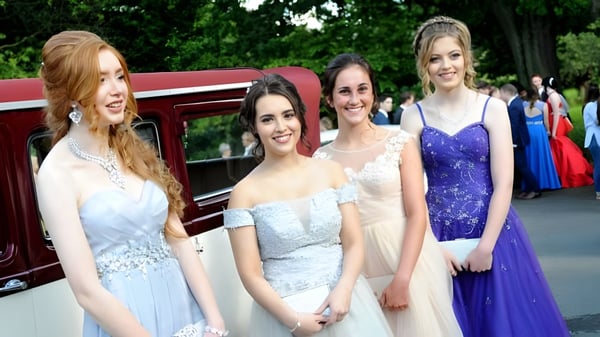 Cuatro alumnas de la Sacred Heart Secondary School Clonakilty están vestidas de forma formal frente a un auto antiguo rojo bajo los árboles.
